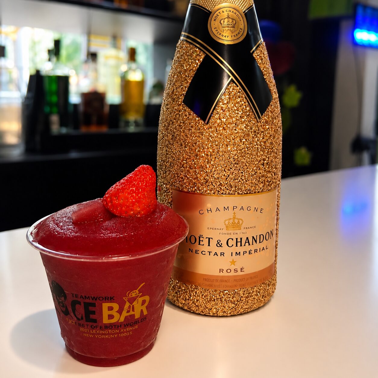 A frozen strawberry drink in a Teamwork Ice Bar cup beside a gold champagne bottle