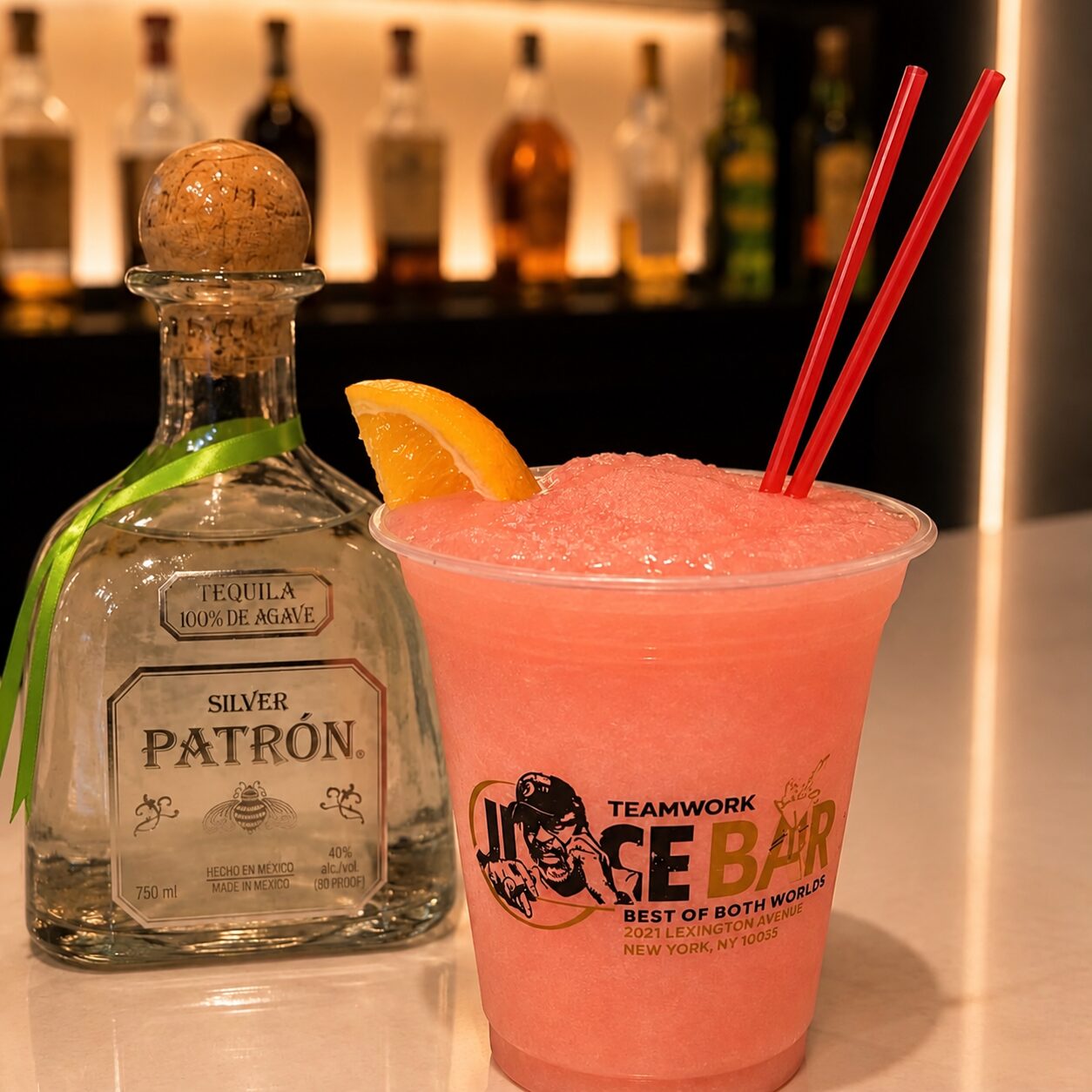 A frozen pink drink with two straws beside a tequila bottle on the bar