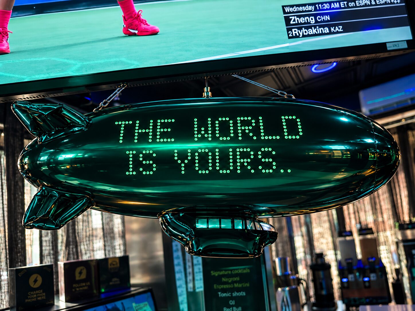 A green blimp balloon lit with the words The World Is Yours, hanging under a screen at the bar