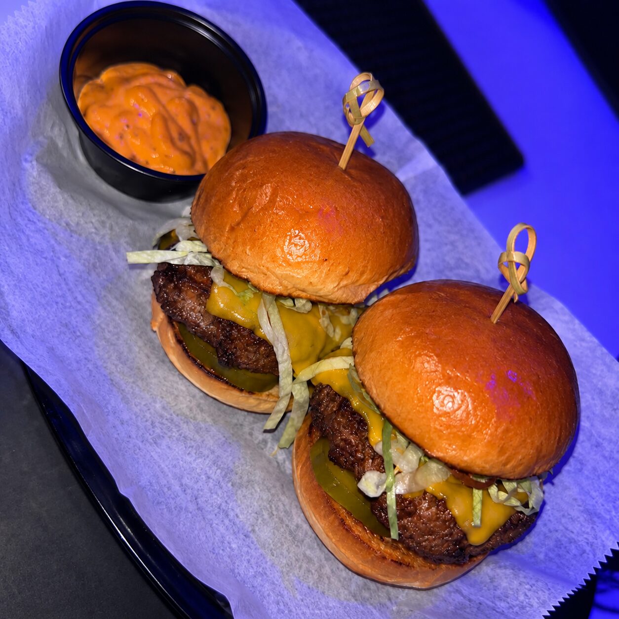 Two cheeseburger sliders on wax paper with a cup of house sauce, under the bar's blue light