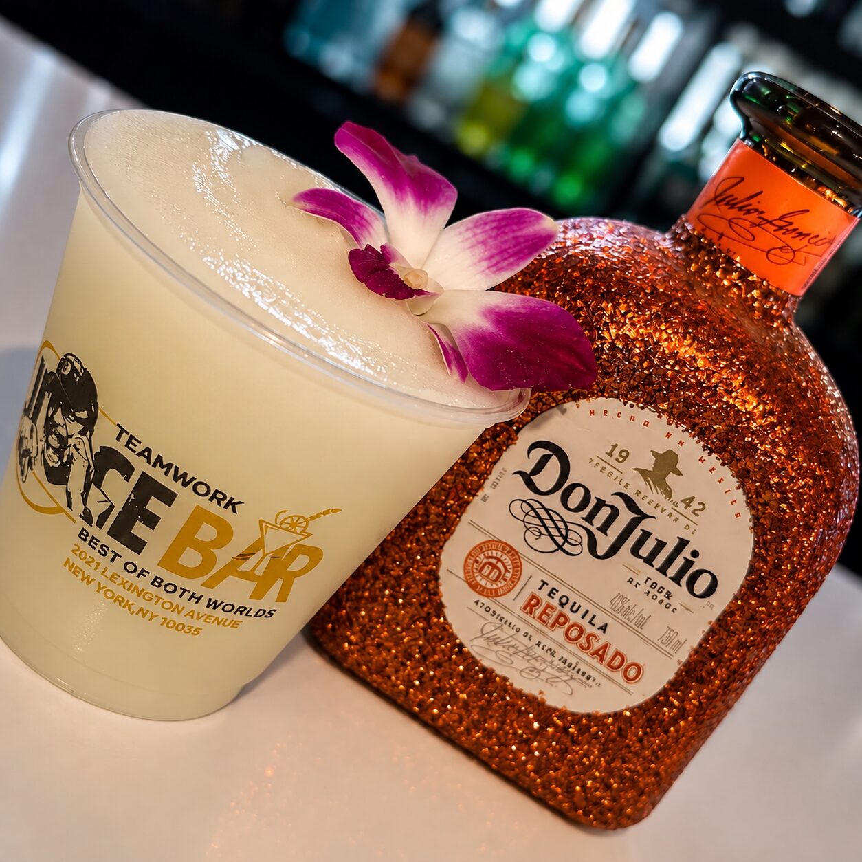 A frozen cocktail in a Teamwork Ice Bar cup, garnished with an orchid, beside a tequila bottle