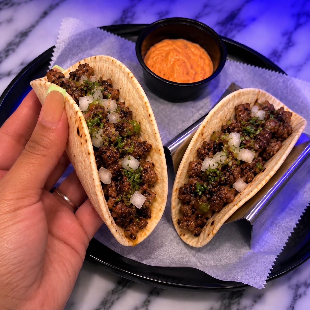 A hand lifting one of two beef tacos topped with onion and cilantro, with a cup of house sauce