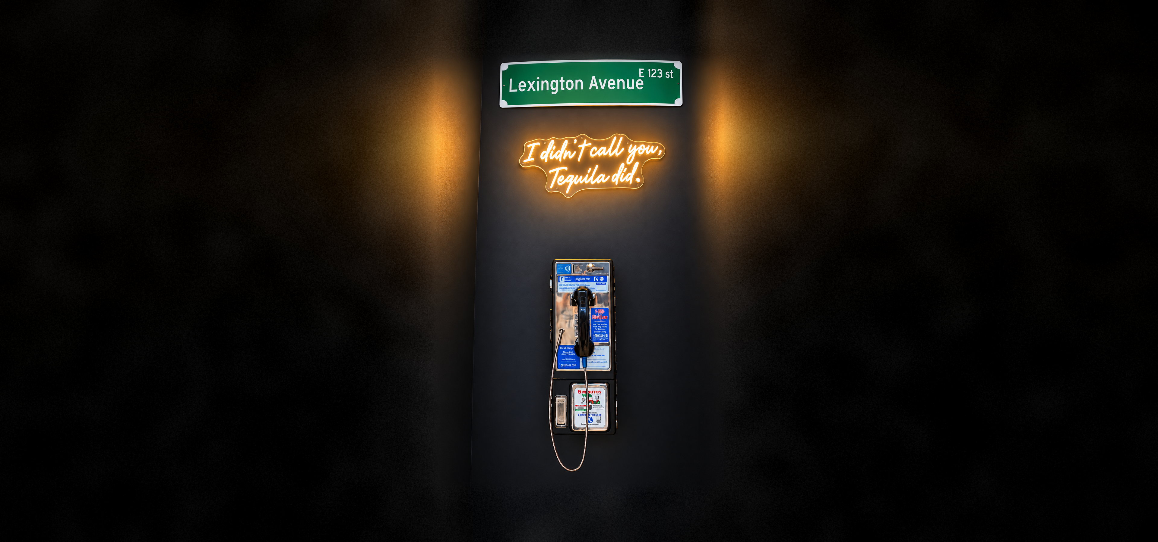 A payphone mounted on a dark wall beneath a neon sign reading "I didn't call you, Tequila did." and a Lexington Avenue and East 123rd Street sign